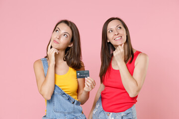 Wall Mural - Two smiling pretty pensive young brunette women friends 20s wearing denim clothes hold credit bank card put hands prop up on chins looking up isolated on pastel pink colour background studio portrait.