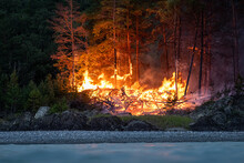 Intense Flames From A Massive Forest Fire At Night. The Flames Light Up At Night As They Rage Through The Forests, Against The Backdrop Of The Mountains And The River