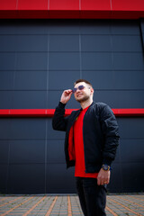 Wall Mural - Young smiling european man in casual wear, red t-shirt, black jacket on a city street against a gray wall background. Stylish modern guy