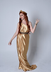 Poster - Full length portrait of girl with red hair wearing long grecian toga and golden wreath. Standing pose iisolated against a grey studio background.