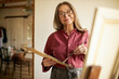 © shurkin_son - Long-haired middle aged female artist in spectacles standing in front of canvas holding brush and palette painting. Beautiful mature woman artist working in her studio. Creativity and inspiration