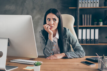 Photo of worried horrified girl sit desk work remote computer bite finger nails feel fear about company crisis bankrupt job loss wear blazer jacket suit in workplace workstation
