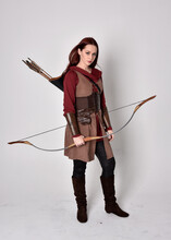 Full Length Portrait Of Girl With Red Hair Wearing  Brown Medieval Archer Costume.. Standing Pose Holding A Bow And Arrow,  Isolated Against A Grey Studio Background.
