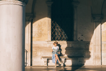 Wall Mural - Cheerful young female tourist holding map and sitting on bench at building