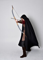 Sticker - Full length portrait of girl with red hair wearing medieval archer costume with black cloak. Standing pose with back to the camera holding a bow and arrow,  isolated against a grey studio background.