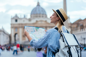 Caucasian traveller with touristic backpack enjoying sightseeing time for exploring Vatican city and ancient St Peter Square using navigation paper map, hipster girl enjoying summer solo vacations