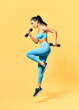 © GVS - Sporty woman in sportswear jumping with dumbbells on yellow background