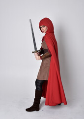 Poster - Full length portrait of girl wearing medieval costume and red cloak. Standing pose holding a sword,  isolated against a grey studio background.