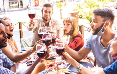 Wall Mural - Friends toasting red wine at restaurant bar with face masks - New normal friendship concept with happy people having fun together on sunny day - Bright filter with focus on right blond woman