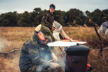 Man hunter cooking on fire outdoors on field kitchen in camping site after hiking    