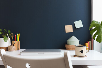 Home office with laptop closed, office supplies, mug, notes, books and dark blue wall. Homeschooling concept. Trendy, creative workspace.