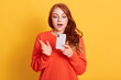 © sementsova321 - Stunned European woman being in shock, reads terrible news from cellular gadget, has wavy red hair, wears orange sweater, poses against yellow wall.