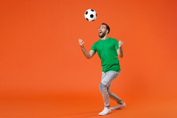 Wall Mural - Full length portrait excited young man football fan in green t-shirt cheer up support favorite team throwing soccer ball isolated on orange background studio. People sport leisure lifestyle concept.