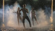 Professional Dancers Perform Salsa Elements In A Dark Smoky Studio. Silhouettes Of Three Bodies In The Rain Moving In Slow Motion.