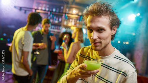 Portrait of caucasian young man looking at camera while posing with a cocktail in his hand and friends chatting, having drinks at the bar counter in the background