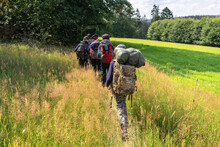 Scouts Or Tourists Go With Big Backpacks On The Green Forest Trail On Sunny Summer Day. Active Healthy Lifestyle People With Rucksacks. Hiking Trekking Scouting. Green Forest Background.