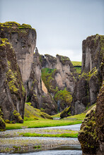 Landscape Vertical View Of Canyon In Fjadrargljufur, Iceland With Large Cliffs And River Water By Green Grass