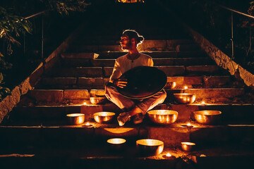 silhouette of a man playing handpan surrounded by Tibetan singing bowls and lighted candles sitting on the steps of the night city. Noise effect on the film