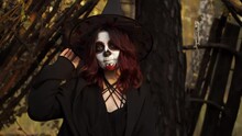 Witch In A Hat With Santa Muerte Makeup Sends Air Kiss In Camera. Portrait Of Woman Sitting In Hut In Forest. A Girl A Halloween Magician Costume Flirts. The Conception Making Witchcraft Of The Woods.