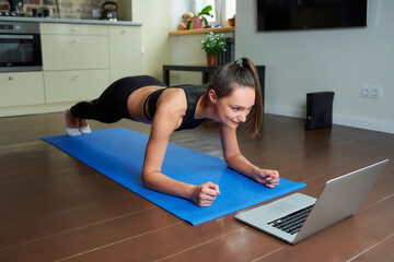 A sporty smiling girl in a black workout tight suit is doing plank watching an online video on a laptop. A female coach conducting a remote fitness class on the blue yoga mat at home.