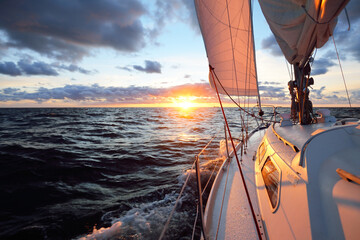 Wall Mural - Yacht sailing in an open sea at sunset. Close-up view of the deck, mast and sails. Clear sky after the rain, dramatic glowing clouds, golden sunlight, waves and water splashes, cyclone. Epic seascape