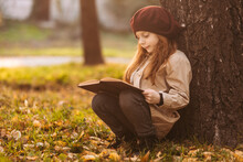 Cute Little Girl Reading A Book Sitting Under A Tree In The Park In Autumn. Learning Concept. Retro Style. Children's Fashion.