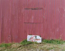 Painted Red Arrow On Piece Of Discarded Wood, In Front Of Old Painted Red Warehouse