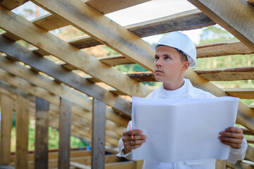 Young male engineer looking at construction work. Foreman in white clothes inspect constructing area. Architect holding blueprint of wooden frame. Wearing helmet for safety. Labor day concept.