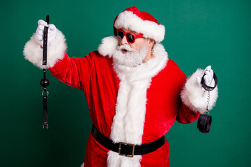 Wall Mural - Portrait of his he nice attractive cheery funny Santa grandfather holding in hands choosing whip tools toys equipment passion erotica game theme party isolated over green color background