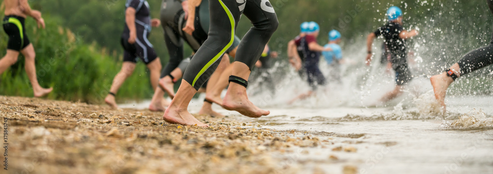 Legs of athletes in wetsuits running into a lake at a triathlon competition - obrazy, fototapety, plakaty 