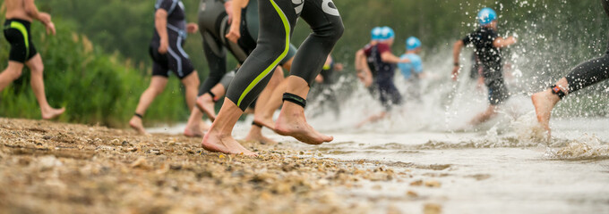 Wall Mural - Legs of athletes in wetsuits running into a lake at a triathlon competition