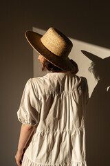 Wall Mural - Young pretty woman in straw hat and white dress / sundress. Sunlight shadow on the wall. Minimal fashion design concept.