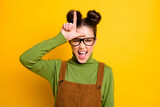 Photo of crazy youngster lady two buns bad person student grimacing showing loser symbol forehead bully classmate wear specs green pullover brown overall isolated yellow color background