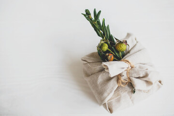 Zero waste christmas holidays. Stylish christmas gift wrapped in linen fabric and decorated with natural green branch on white rustic table background, space for text. .