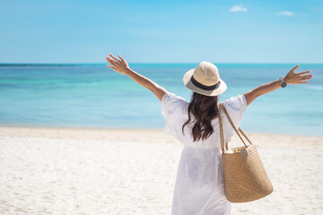 Happy traveler woman in white dress and hat enjoy beautiful sea view, young woman standing on sand and looking ocean at tropical beach. Freedom, relaxing, vacation holiday and summer travel concept