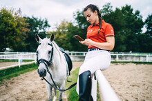 Female Jockey Using Smartphone Sitting On Fence Near Horse