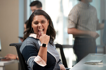 Young woman sitting at computer in office looking to the camera making funny face, co-workers having discussion around dry erase board in background 