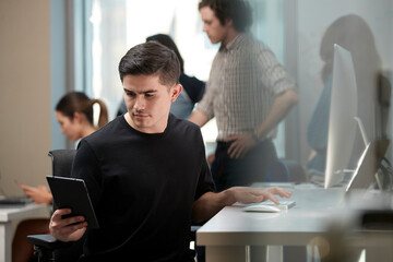 Young man multi-tasking by sitting at computer in office and checking tablet, co-workers having discussion around dry erase board in background 