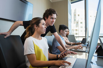 Young woman using computer in office typing on keyboard and checking mobile phone, showing male co-worker something on her screen with co-workers working in background 