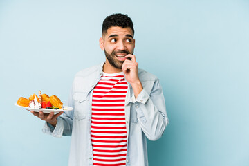 Wall Mural - Young latin man holding a waffle isolated relaxed thinking about something looking at a copy space.