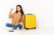 © Asier - Young chinese traveler woman sittting on the floor with a suitcase isolated holding something little with forefingers, smiling and confident.