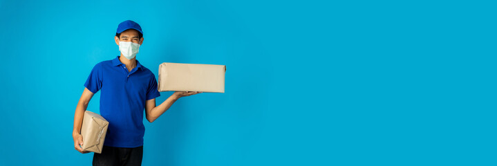 Wall Mural - Asian delivery man wearing mask holding parcel box on blue background in studio