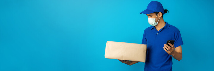 Asian delivery man wearing mask holding and check parcel box and phone on blue background in studio