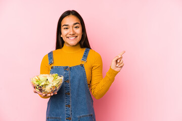 Young asian woman holding a salad isolated smiling and pointing aside, showing something at blank space.