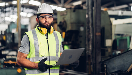 Young caucasian engineering man in Hard Hat Wearing Safety Jacket working on laptop computer in Heavy Industry Manufacturing Facility.