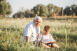 © Maria - Mid shot of grandfather and his grandson while reading a book together in meadow Small boy making first steps in read.
