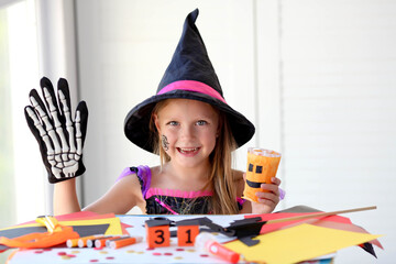 A little girl in a witch costume looks at the camera and laughs, holding a glass pumpkin and a skeleton glove in her hands.