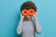 Lovely romantic young woman with flowers in front of eyes, keeps lips rounded, holds orange gerbera daisy, dressed in grey turtleneck, blue background, has spring mood, enjoys pleasant smell