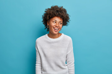 Wall Mural - Joyful dark skinned young woman with broad smiles, perfect teeth, feels carefree and enthusiastic, looks happily aside, stands entertained, wears casual white jumper, isolated on blue background