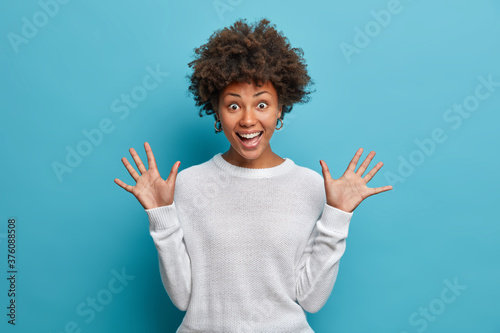 Fotomural Amused pretty girl with curly Afro hair, raises palms, has cheerful expression, smiles broadly, sees something funny, wears white sweater, isolated on blue background
